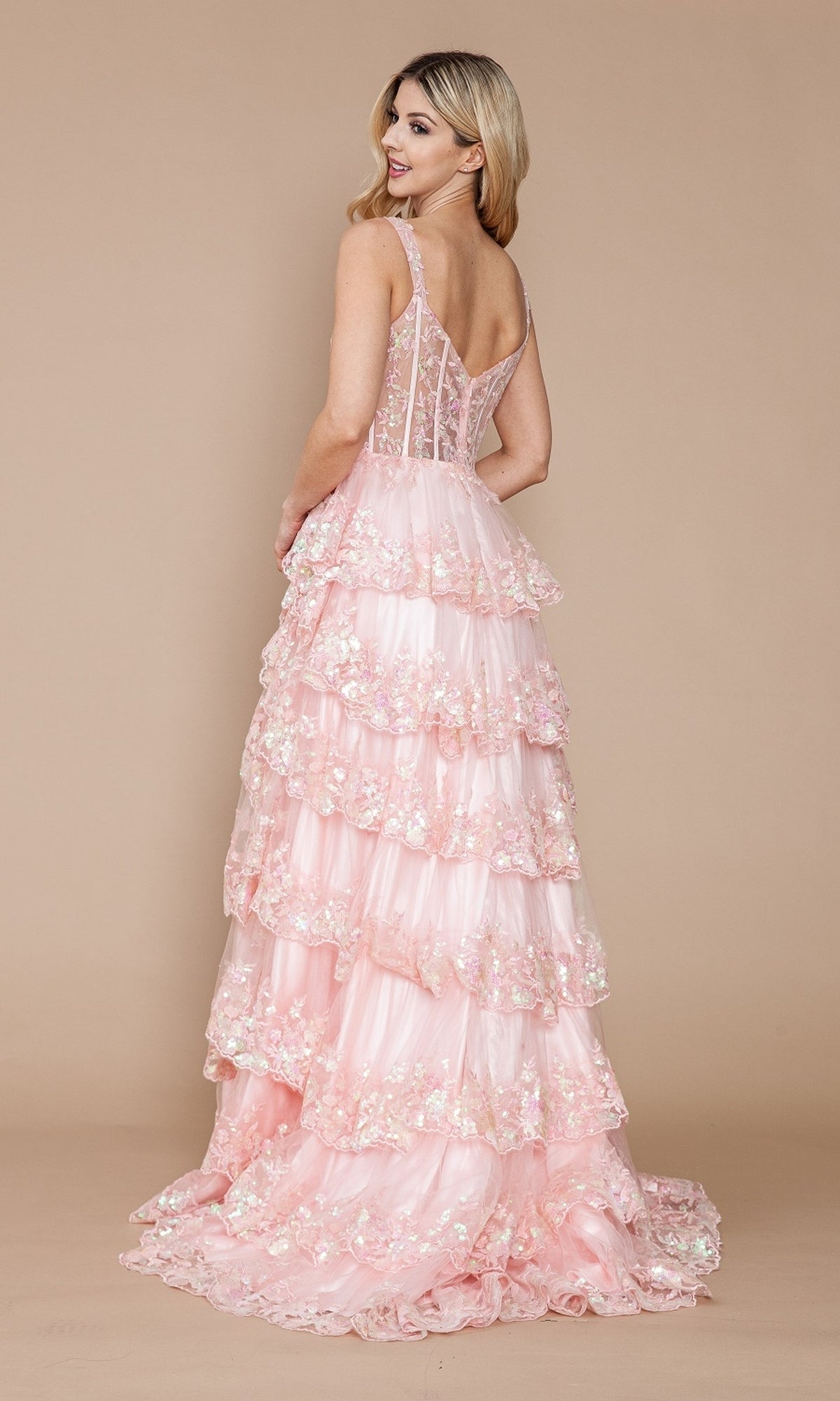 A woman poses against a plain beige background in the Sheer-Corset Long Ruffled Prom Dress: Poly USA 9410—a floor-length, light pink dress featuring floral embellishments, ruffles, and a sheer corset bodice.